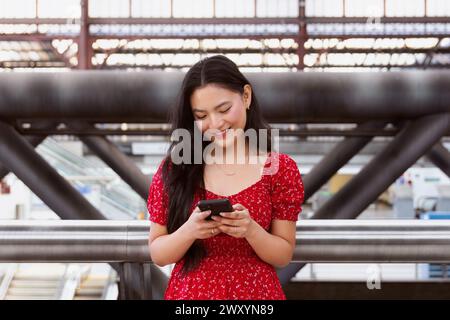 Une jeune femme joyeuse dans une robe rouge se tient debout avec son smartphone dans une gare animée, avec des détails architecturaux et la vie urbaine dans le backgro Banque D'Images