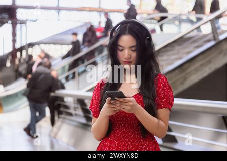 Une jeune femme en robe rouge se concentre sur son smartphone, portant des écouteurs, dans une gare animée Banque D'Images