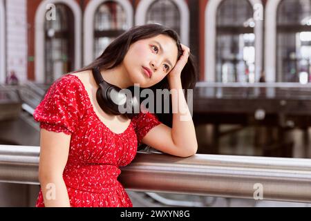 Une jeune femme pensive dans une robe rouge avec des écouteurs autour du cou s'appuie sur une balustrade dans un bâtiment moderne Banque D'Images