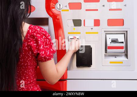 Une femme aux cheveux noirs vue de derrière utilise un distributeur automatique de billets, tenant une carte et utilisant le clavier Banque D'Images