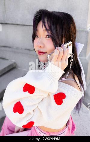 Une fille chinoise Gen-Z branchée avec une expression ludique parle sur son smartphone, portant un pull blanc avec des coeurs rouges et des accessoires élégants Banque D'Images