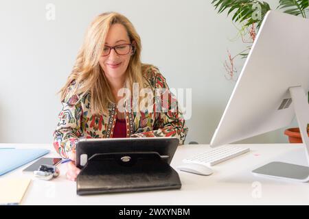 Une femme d'affaires blonde professionnelle travaillant attentivement à son bureau avec une tablette et un ordinateur Banque D'Images