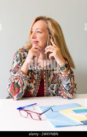 Une femme blonde professionnelle s'engage dans une conversation téléphonique, regardant contemplative, avec des accessoires de bureau sur son bureau Banque D'Images