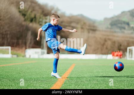 Un jeune athlète en uniforme bleu pratique le soccer en frappant une balle rouge et bleue sur un terrain ensoleillé Banque D'Images