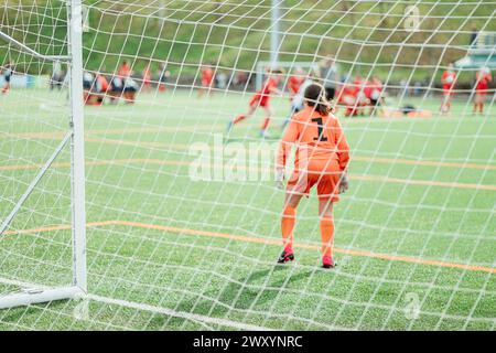 Un jeune joueur de football se tient dans le but, enfilant un uniforme orange et des gants, regardant attentivement un match sur un terrain de gazon artificiel vert Banque D'Images