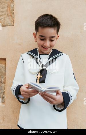 Jeune garçon souriant engagé dans la lecture d'un livre de prières, portant sa première tenue de communion, avec un collier en croix Banque D'Images