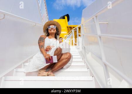 Femme souriante en robe blanche et chapeau de soleil se trouve sur l'escalier d'un navire, tenant un passeport, profitant de son escapade de week-end. Banque D'Images