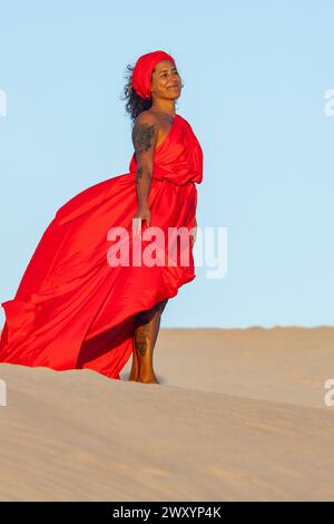 Une femme contente dans une robe rouge fluide et un foulard se tient sur les dunes du désert, regardant loin de la caméra avec une expression sereine. Banque D'Images