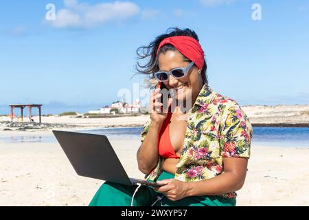 Nomade numérique joyeux s'engageant dans un appel d'affaires tout en tenant un ordinateur portable sur une plage ensoleillée, mélangeant travail et cadre détendu Banque D'Images