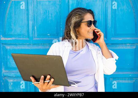 Une femme souriante en lunettes de soleil se tient contre une porte bleue, tenant un ordinateur portable et parlant sur un téléphone portable, représentant un style de vie nomade numérique. Banque D'Images