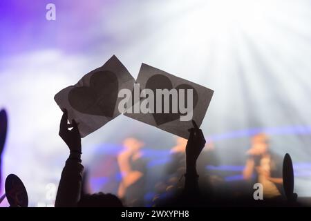 Mains anonymes aux silhouettes tenant des découpes en forme de cœur lors d'un concert animé, capturant un moment d'unité et d'adoration des fans dans une ambiance de scène éclatante Banque D'Images