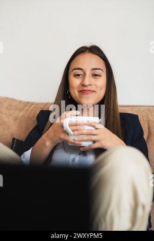 Une jeune femme d'affaires à l'aise sur un canapé, tient une tasse à café avec un sourire content Banque D'Images