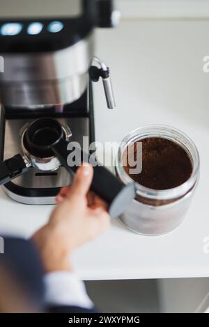 La main d'une personne tenant un porte-filtre à côté d'un pot de café moulu et d'une machine à expresso Banque D'Images
