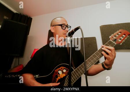 Un homme jouant d'une guitare acoustique chante dans un microphone, regardant loin de la caméra dans un studio de musique. Banque D'Images