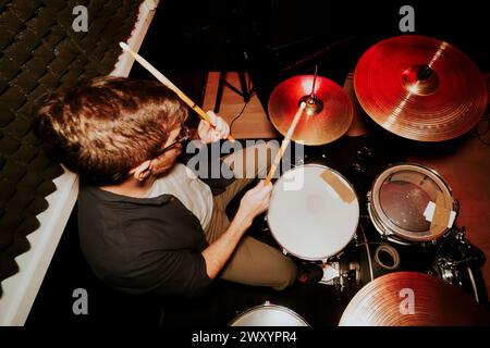 From Above batteur masculin regardant loin se concentre intentionnellement sur son set de batterie pendant une performance, avec des cymbales et des baguettes de batterie en vue. Banque D'Images