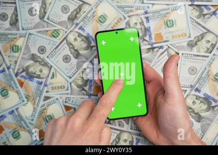Main tenant smartphone avec écran vert sur pile de billets de dollars, vue de dessus. Concept de gains en ligne Banque D'Images