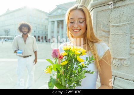 Femme heureuse avec un bouquet de fleurs, son partenaire derrière avec une boîte est sur le point de proposer Banque D'Images