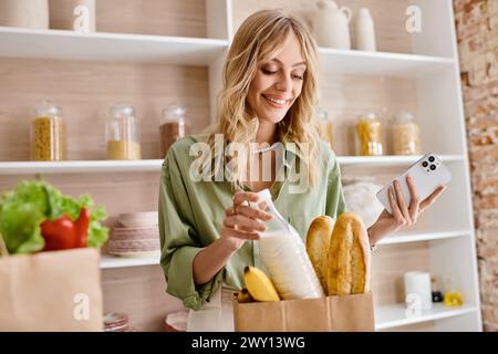 Une femme debout dans une cuisine tenant un sac de pain et un téléphone portable. Banque D'Images