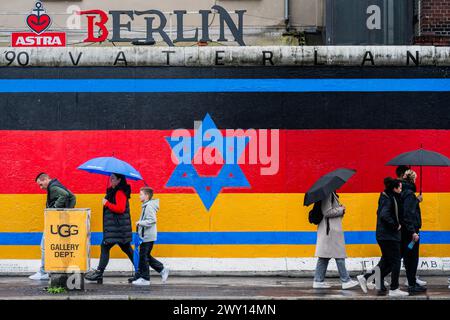 Berlin, Allemagne. 3 avril 2024. Un drapeau israélien est mélangé avec le drapeau allemand alors que les gens passent avec des parapluies et des imperméables par temps pluvieux - la East Side Gallery à Berlin. Un projet de graffiti conservé par 118 artistes couvrant une partie du mur de Berlin et célébrant sa disparition. Crédit : Guy Bell/Alamy Live News Banque D'Images