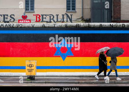 Berlin, Allemagne. 3 avril 2024. Un drapeau israélien est mélangé avec le drapeau allemand alors que les gens passent avec des parapluies et des imperméables par temps pluvieux - la East Side Gallery à Berlin. Un projet de graffiti conservé par 118 artistes couvrant une partie du mur de Berlin et célébrant sa disparition. Crédit : Guy Bell/Alamy Live News Banque D'Images