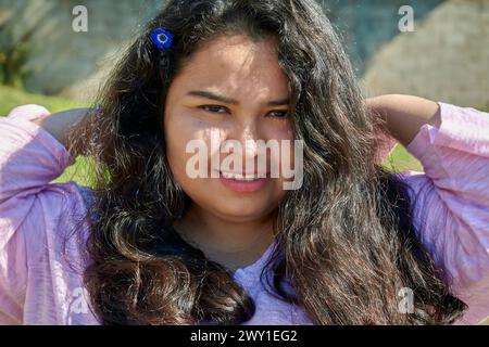 Portrait d'une jeune femme latino-américaine un jour d'été. Elle a de longs cheveux noirs. Elle sourit à la caméra. Elle a une fleur bleue dans les cheveux. Banque D'Images