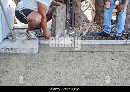 Installation d'un nouveau pavé ou plancher à l'extérieur de grands carreaux de béton, gros plan sur le travailleur masculin préparant le bloc de pierre pour s'adapter sur le sable et le gravier BA Banque D'Images