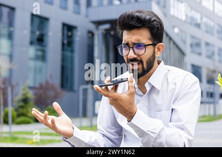 Inquiet et contrarié jeune Indien en chemise blanche et lunettes assis près du bureau de travail et parlant au téléphone par haut-parleur, et enregistrant un message vocal. Banque D'Images