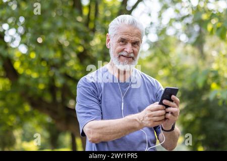 Portrait en gros plan d'un homme âgé aux cheveux gris debout dans un parc à l'extérieur, portant des écouteurs et tenant un téléphone, faisant du sport et marchant, souriant à la caméra. Banque D'Images