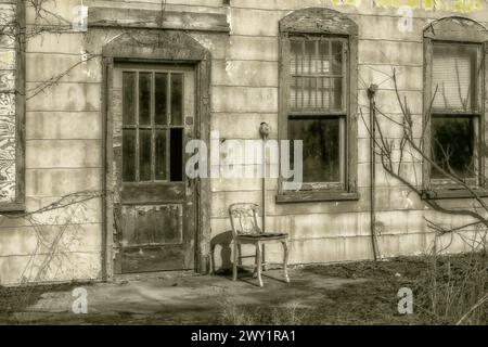 Gros plan noir et blanc d'un vieux bâtiment abandonné où une chaise en bois se trouve à côté de la porte avec des vitres brisées Banque D'Images