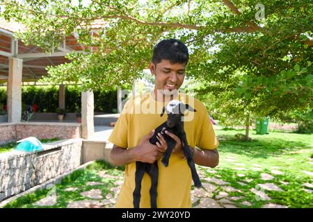 Un fermier indien bienveillant en vêtements traditionnels tient doucement un bébé chèvre noir câlin dans une ferme ensoleillée. Banque D'Images