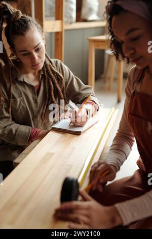 Jeune femme sérieuse avec des dreadlocks regardant son collègue avec un ruban à mesurer prenant des mesures de banc ou de planche en bois Banque D'Images