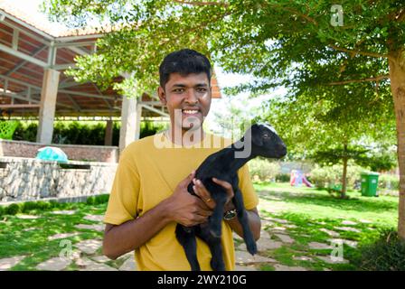 Un fermier indien au visage gentil en vêtements traditionnels tient doucement un gosse de chèvre noir moelleux dans ses bras sur une ferme lumineuse et ensoleillée Banque D'Images
