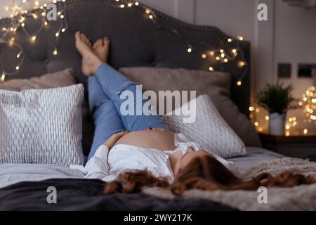 Belle femme enceinte rousse d'âge moyen attendant un bébé allongé sur le lit détendu avec les jambes vers le haut Banque D'Images