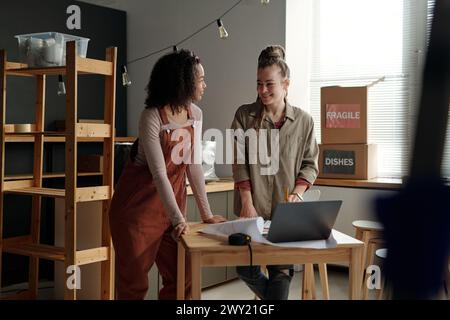 Deux jeunes femmes entrepreneurs heureux ou designers d'intérieur debout près de la table devant l'ordinateur portable et discutant de certaines questions de travail Banque D'Images