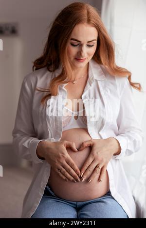 Belle femme enceinte rousse d'âge moyen attendant un bébé avec les mains formant un cœur sur le ventre à la maison Banque D'Images