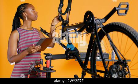 Femme afro-américaine faisant le service de vélo dans un atelier de réparation de fond de studio, regardant sur la liste d'entretien de tablette. Technicien vérifiant les composants de bicyclette qui ont besoin d'être réparés, caméra B. Banque D'Images