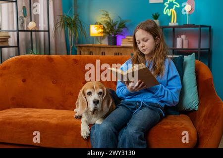 Fille preteen heureuse lisant un livre intéressant, tournant les pages souriantes appréciant la littérature, prenant un repos sur un canapé confortable. Portrait d'enfant heureux à la maison appartement salon sur canapé avec chien de compagnie Banque D'Images