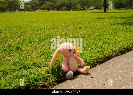 Lapin farci rose assis dans l'herbe verte à côté du trottoir. Pieds violets et jaunes avec des arcs roses jaunes sur la tête. Oreilles pendantes. Lapin de Pâques Banque D'Images