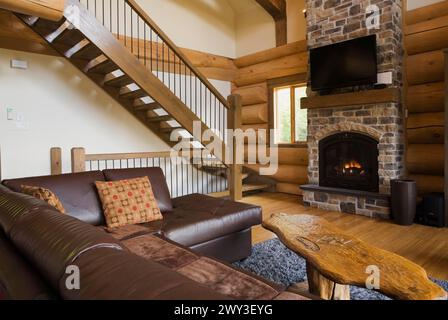 Canapé en l en cuir marron et table basse en bois devant un foyer au propane en pierre naturelle brune et bronzée nuancée à côté d'escaliers en bois Banque D'Images