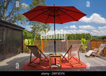 Belvédère en filet noir avec parasol en toile rouge et chaises pliantes en toile en bois sur tapis rouge sur terrasse en planche de bois gris avec balustrade donnant sur le lac Banque D'Images