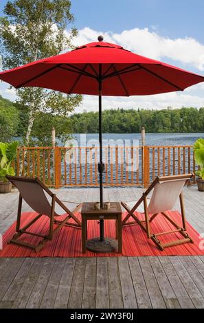 Parasol en toile rouge et chaises en toile pliantes en bois sur tapis rouge sur pont de planche en bois gris avec balustrade donnant sur le lac avec forêt verte dedans Banque D'Images