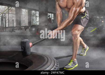 Homme musclé sportif frappant pneu de roue avec traîneau de marteau dans la salle de gym. Entraînement sportif Banque D'Images