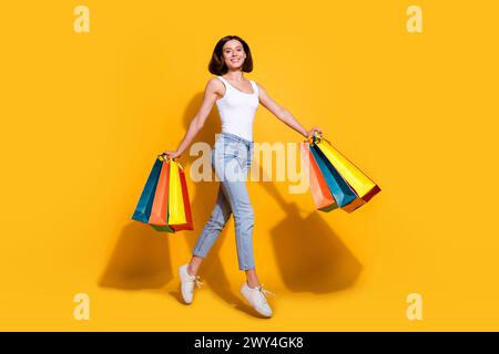 Photo du corps entier de jeune fille attrayante tenir des sacs à provisions en cours d'exécution porter une tenue blanche à la mode isolée sur fond de couleur jaune Banque D'Images