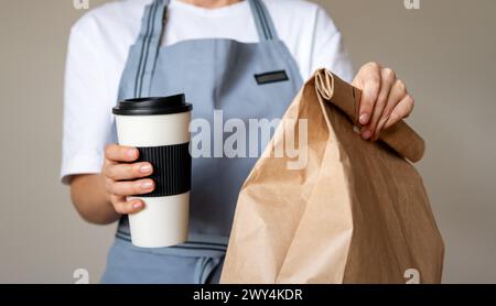Repas à emporter entre les mains de la serveuse. Tasse à café de voyage et sac d'emballage en papier brun avec de la nourriture à l'intérieur. Banque D'Images