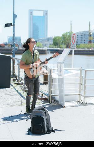 Image verticale d'un jeune homme latino d'ethnie vénézuélienne busker avec de longs cheveux et des lunettes, travaillant à la guitare et chantant à Puerto Madero Arge Banque D'Images