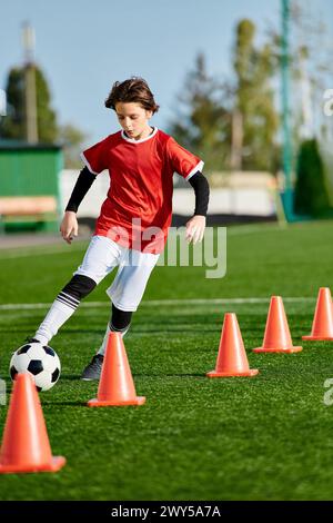 Un jeune garçon lance énergiquement un ballon de football autour de cônes orange, montrant son agilité et son habileté sur le terrain. Banque D'Images