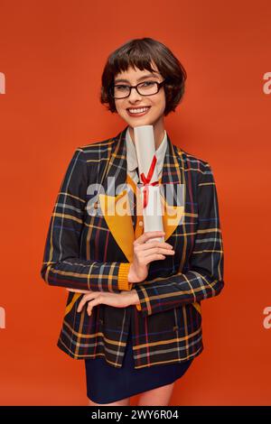 fille collégiale souriante en uniforme et lunettes tenant son diplôme sur fond orange Banque D'Images