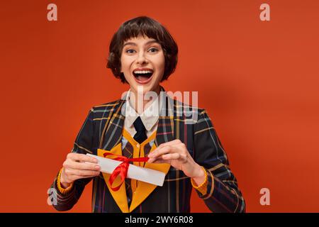 portrait d'une fille d'université excitée en uniforme à carreaux tenant son diplôme sur fond orange Banque D'Images