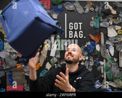 Mainz, Allemagne. 04th Apr, 2024. Benjamin Mandos, fondateur de GOT Bag, basé à Mayence, attrape un sac à dos devant un mur avec des déchets plastiques collés autour du logo de la marque. Fondée en 2016, la start-up utilise pour ses sacs à dos et valises des tissus fabriqués à partir de plastique recyclé provenant de la mer ou de la mangrove, connu sous le nom de plastique Ocean impact. Crédit : Arne Dedert/dpa/Alamy Live News Banque D'Images
