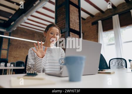 Heureuse femme freelance parlant avec un collègue via un appel vidéo et agitant Salut assis dans le coworking Banque D'Images
